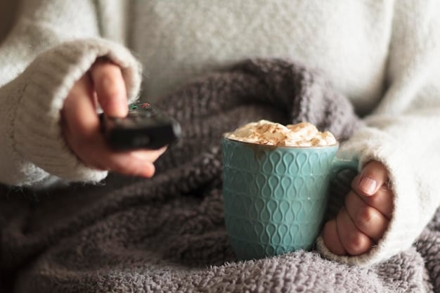 A person sitting comfortably on a couch with a blanket, watching a movie on a large screen TV, with a coffee table in front holding snacks and drinks. The setting emphasizes the comfort and convenience of watching movies at home.