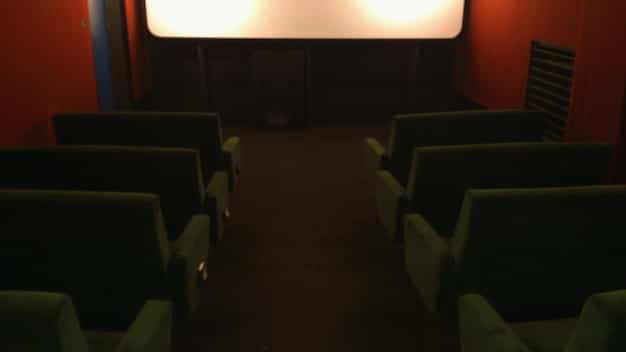 A dimly lit, nearly empty independent movie theater auditorium. Only a few patrons are scattered throughout the seats, watching a movie screen. The atmosphere is quiet and somewhat melancholic.