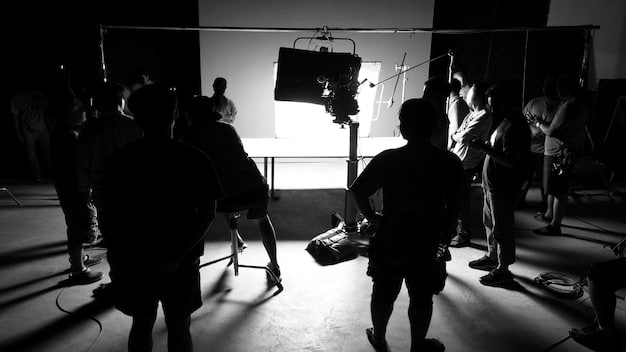 A cinematic shot of a crowded film set. Directors are sitting behind monitors. In the foreground, an actor dressed in a costume. Lighting equipment and crew members surround the scene.