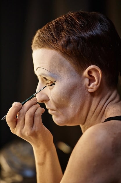 A close-up shot of a makeup artist meticulously applying prosthetic makeup to an actor's face. The focus is on the detail and precision involved, with tools and various makeup products visible. The actor is expressing comfort.
